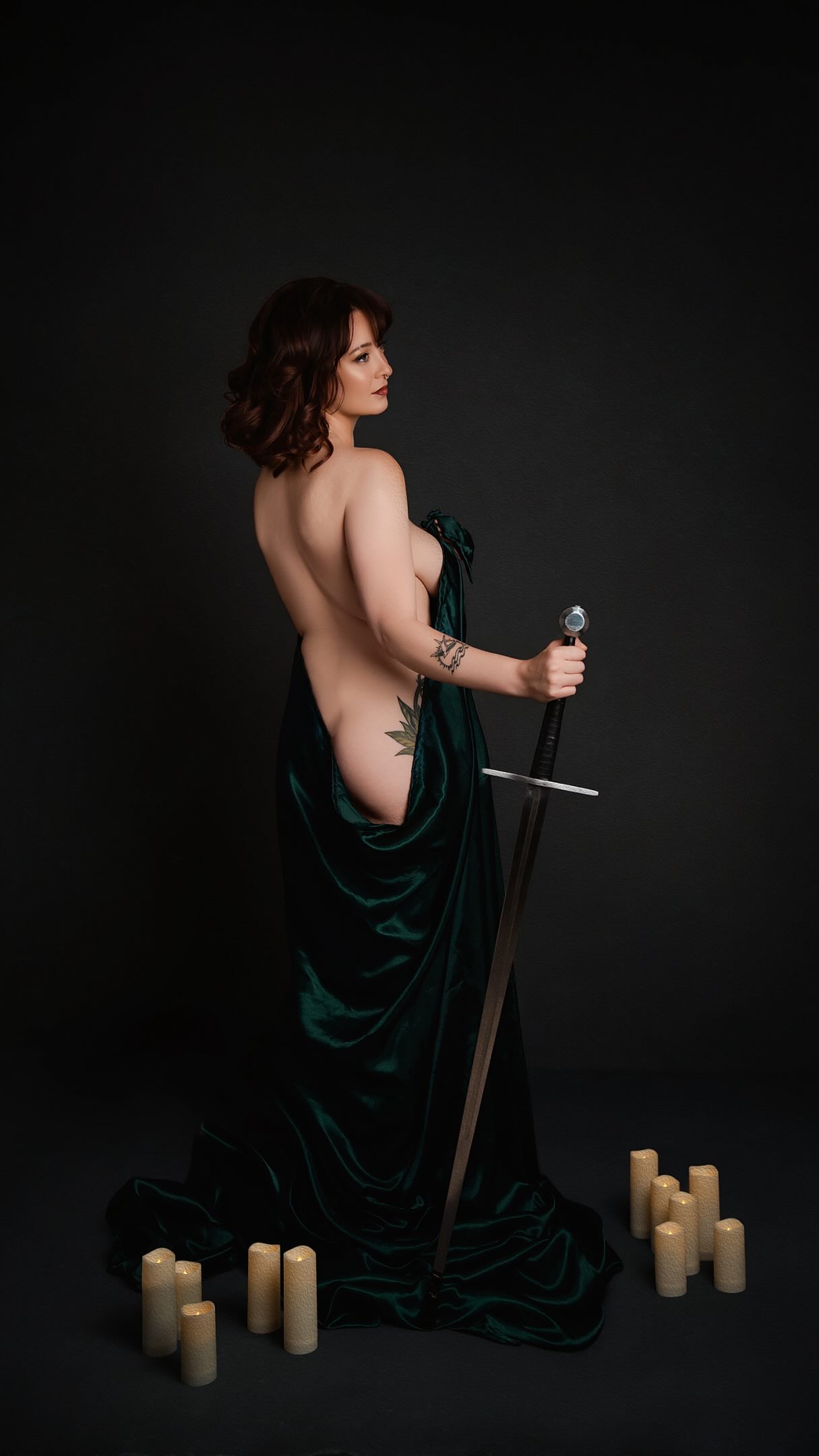 Woman draped in green velvet holding a sword beside candles